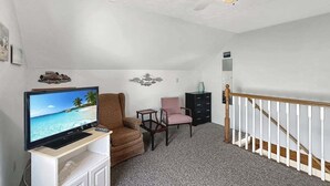 Um quarto com televisão sobre suporte, uma poltrona marrom, uma cadeira rosa, uma mesa pequena e uma escada com corrimão branco.