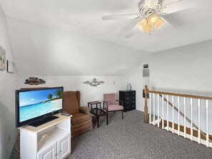 Ein Zimmer mit Fernseher auf einem Schrank, einem braunen Sessel, einem rosa Stuhl, einem kleinen Tisch und einer Treppe mit einer weißen Geländer.