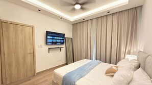Una moderna camera d'albergo con un letto, tende, una ventola a soffitto e un televisore a muro che mostra servizi di streaming.