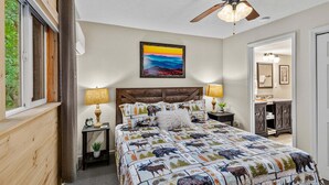 A bedroom with a bed, a ceiling fan, a painting, a nightstand, and a view of the bathroom. - Bearfoot Beech - Game table, fire pit, pets ok, hot tub! (Banner Elk)