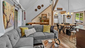 A modern interior with a grey sofa, wooden dining table, and a kitchen area. - Bearfoot Beech - Game table, fire pit, pets ok, hot tub! (Banner Elk)