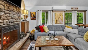 A cozy living room with a stone wall, fireplace, grey sofa, and a view of greenery outside. - Bearfoot Beech - Game table, fire pit, pets ok, hot tub! (Banner Elk)