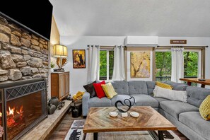 Ein gemütliches Wohnzimmer mit Steinwand, Kamin, grauem Sofa und Blick auf Grünflächen.