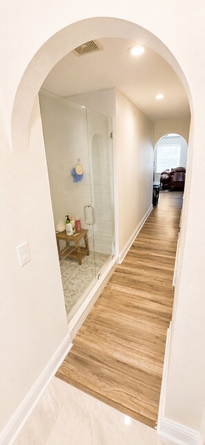 A hallway with a wooden floor, a glass shower enclosure, and a bench with toiletries.
