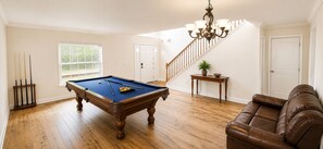 A room with a pool table, a brown leather sofa, a staircase, and a chandelier.