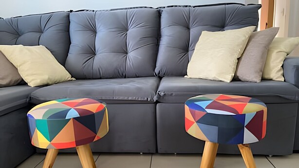 Ein graues Ecksofa mit vielen Kissen und zwei farbenfrohe geometrische Hocker mit Holzbeinen.