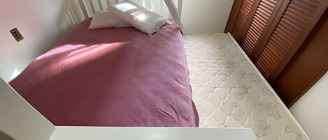 Una habitación pequeña con cama individual, cabecera blanca, colcha rosa y ventana con persianas.