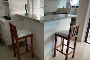 A kitchen with a central island, two wooden bar stools, white cabinetry, and a window providing natural light. - SHB Joia de Pitangueiras lazer completo (Guarujá)