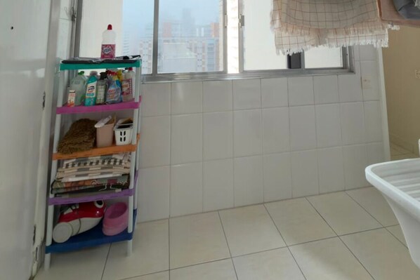 Un baño con piso de baldosas, un estante con productos de limpieza y una ventana con vista a edificios.