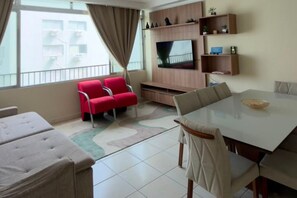A modern living room with a flat-screen TV, red chairs, a dining table, and a balcony with a view. - Fully Furnished Apartment in the Heart of Pitangueiras, Spacious Apartment in Guarujá (Guarujá)