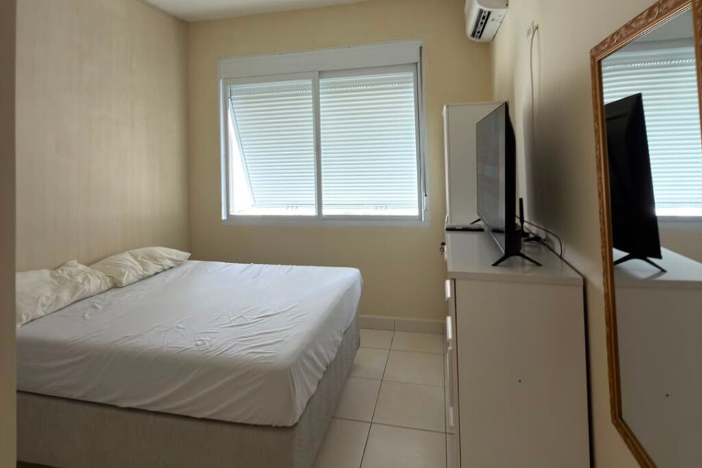 Ein Schlafzimmer mit Bett, Fernseher auf einem Schrank, einem Fenster mit Jalousien und einem Spiegel an der Wand.