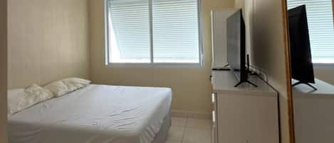 A bedroom with a bed, a television on a stand, a window with blinds, and a mirror on the wall.