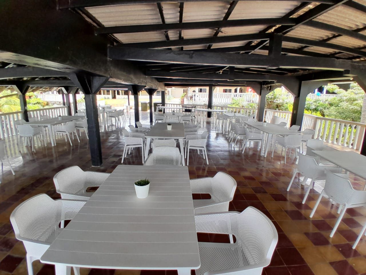 An outdoor covered dining area with multiple tables and chairs, a tiled floor, and a view of buildings in the background.