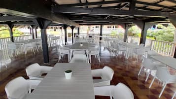 An outdoor covered dining area with multiple tables and chairs, a tiled floor, and a view of buildings in the background.