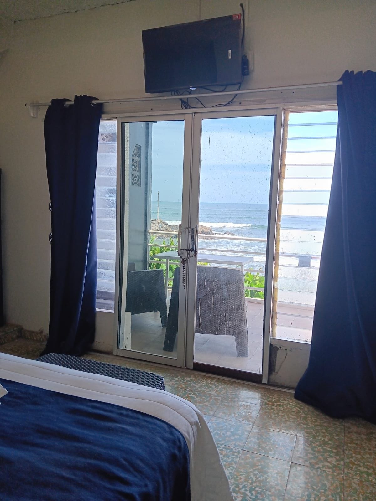 A room with a bed, a television on the wall, and a sliding glass door leading to a balcony with a view of the beach.