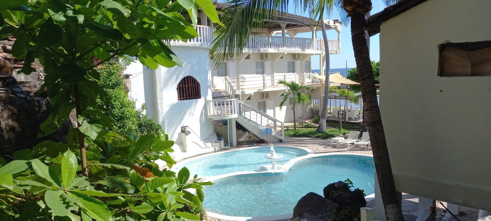 Zona de piscina con una fuente, rodeada de exuberante vegetación y un edificio de varios pisos con balcones.