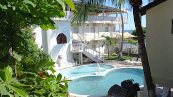 Zona de piscina con una fuente, rodeada de exuberante vegetación y un edificio de varios pisos con balcones.