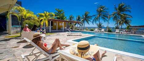 Un espace aménagé au bord de la piscine, avec des chaises longues, une piscine et des palmiers.