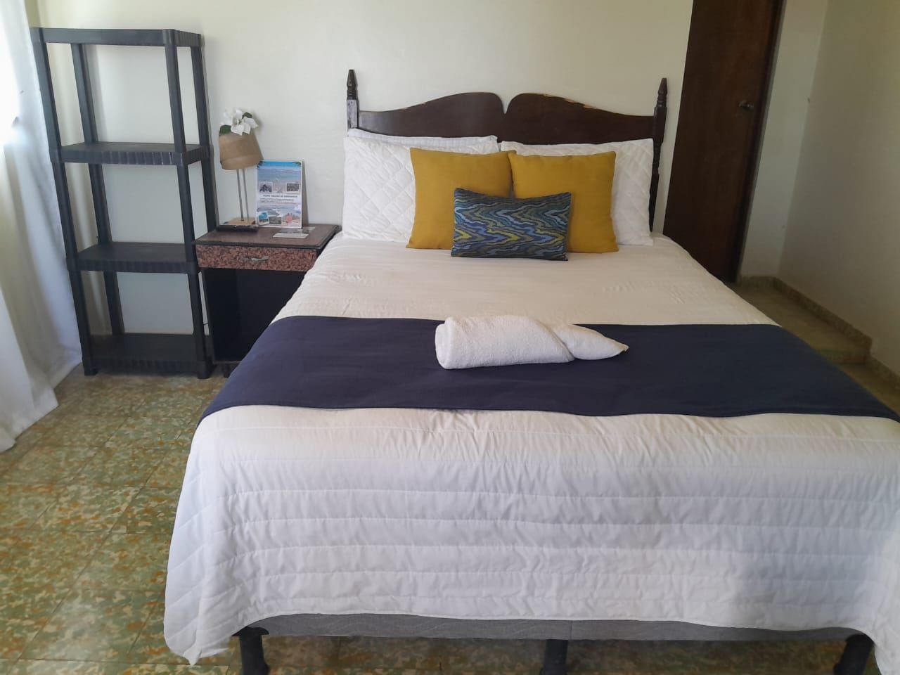 Cama bem arrumada com colcha branca, cobre azul escuro e dois travesseiros, um amarelo e outro com padrão azul e verde.