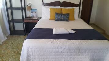 Cama bem arrumada com colcha branca, cobre azul escuro e dois travesseiros, um amarelo e outro com padrão azul e verde.