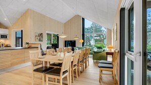 Una cocina moderna con armarios de madera, un comedor con una mesa grande y sillas, y vistas a zonas verdes a través de amplios ventanales.