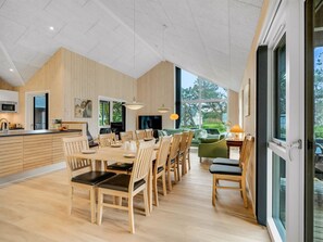 Eine moderne Küche mit Holzausstattung, ein Essbereich mit großem Tisch und Stühlen sowie Blick auf Grünflächen durch große Fenster.