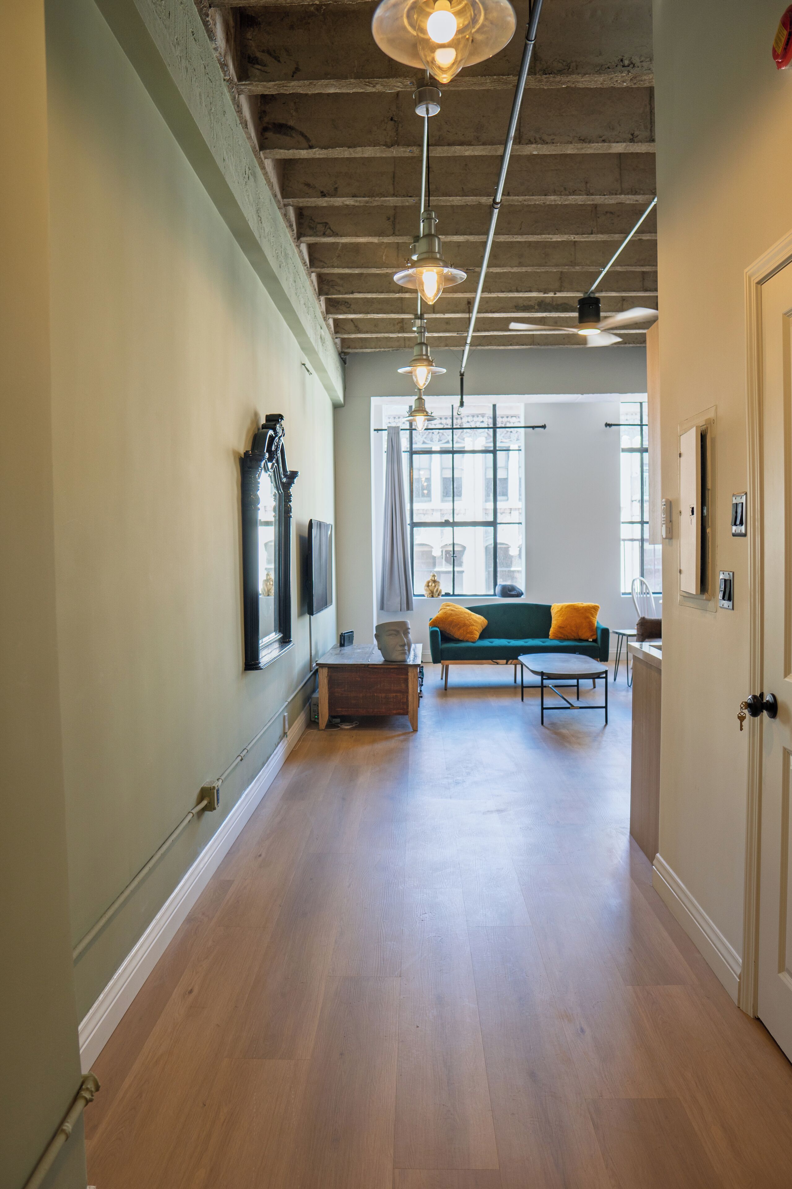 A hallway with wooden flooring, a green sofa with yellow cushions, and a window with curtains.