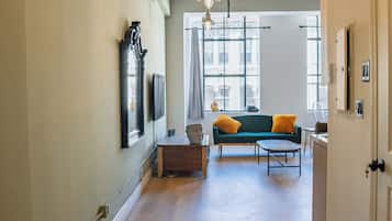 A hallway with wooden flooring, a green sofa with yellow cushions, and a window with curtains.