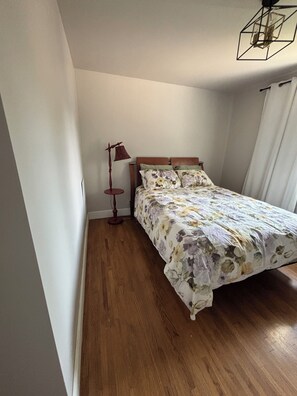 A bedroom with a wooden floor, a bed with floral bedding, a floor lamp, and a geometric ceiling light fixture. - Cozy home away from home in NW Calgary. Great location and has all amenities (Calgary)