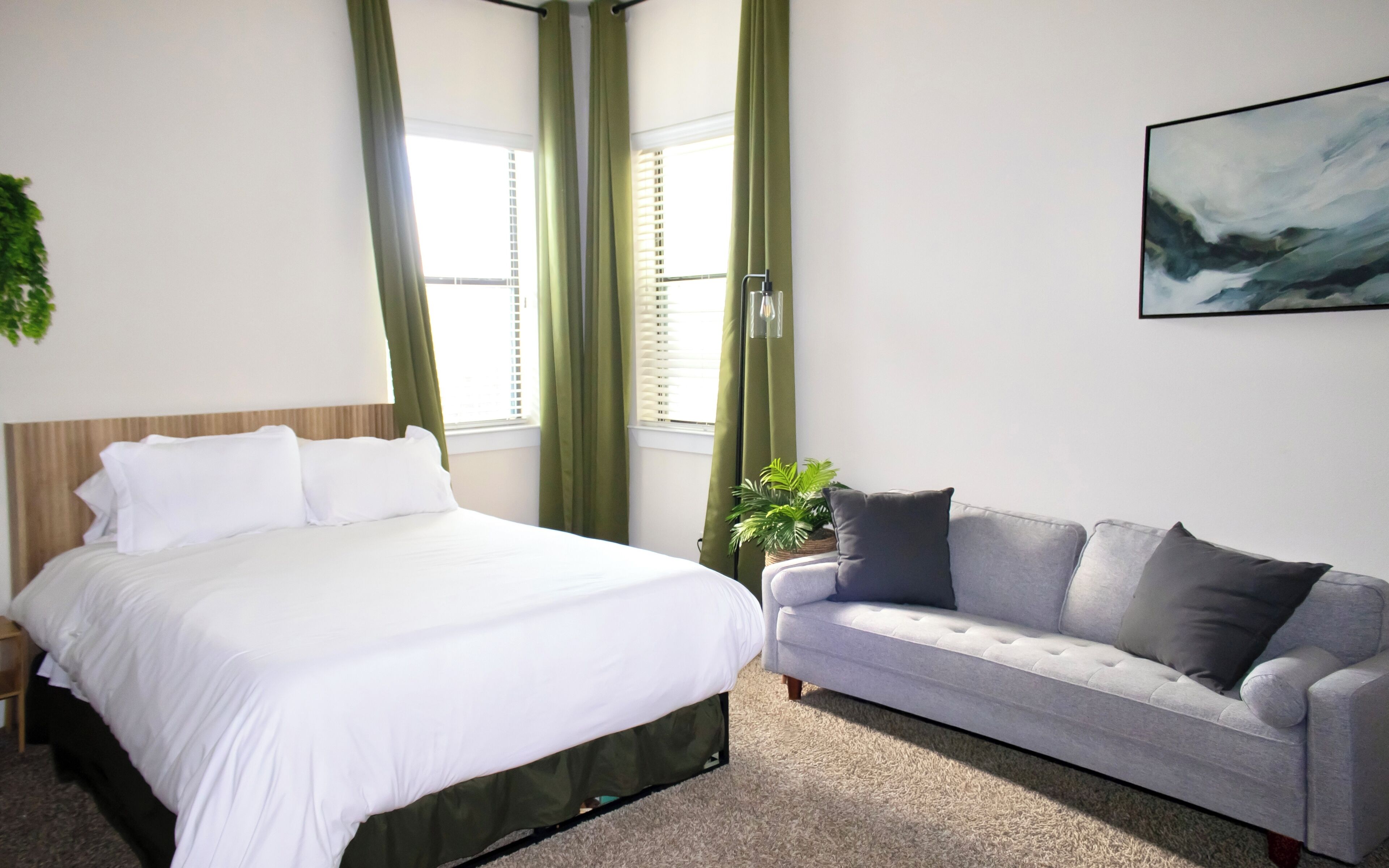 A bedroom with a bed, a grey sofa, a potted plant, and a painting on the wall.