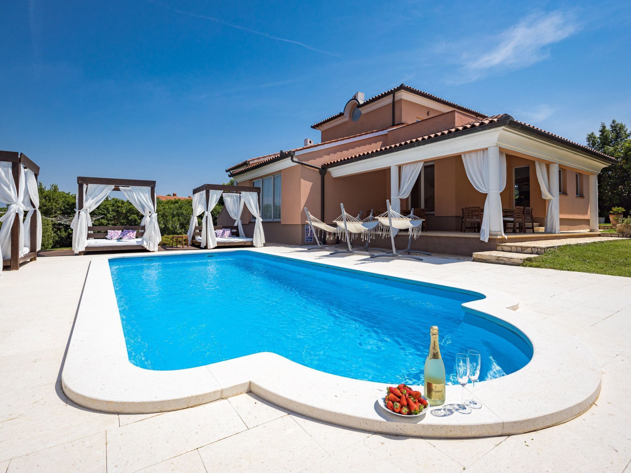 Ein Swimmingpool mit weißer Liegefläche, einem überdachten Sitzbereich und einem Haus mit Ziegeldach.