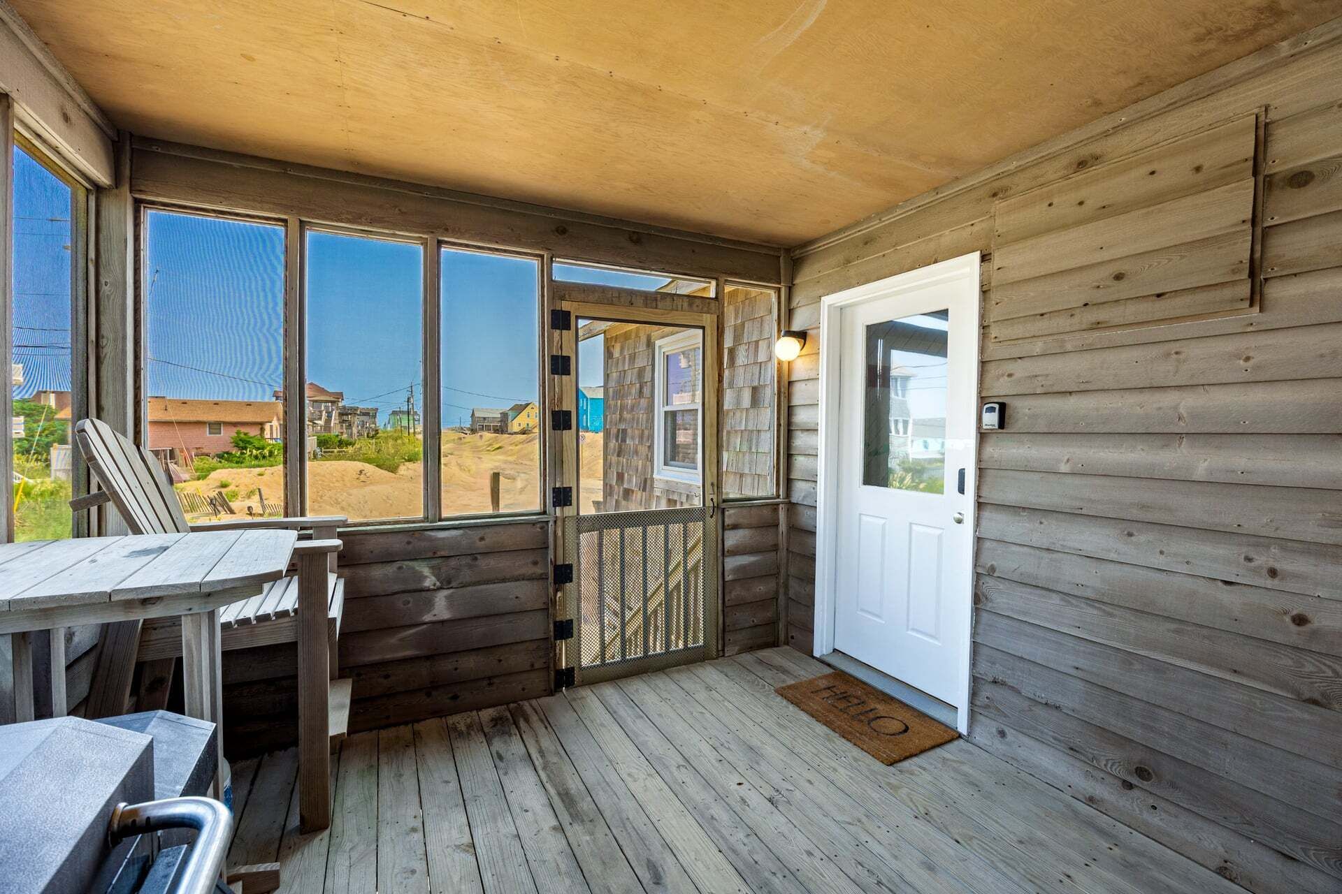 En veranda av trä med ett bord och stolar, en dörr och utsikt över ett sandigt landskap.