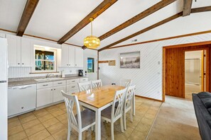 A kitchen with white cabinets, a wooden dining table, and chairs. There is a window with a view of houses, a hanging light fixture, and a door leading to another room. - Saltwater Sunrise: Ocean Views & Beach Access (Rodanthe)