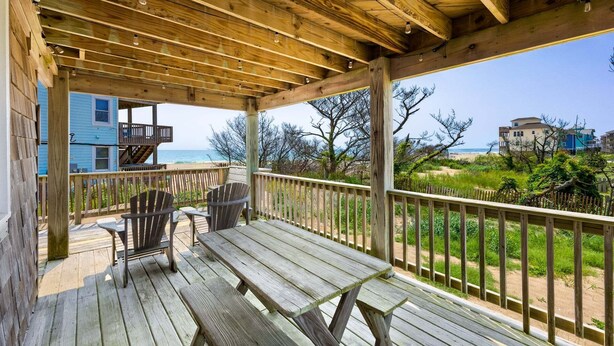 Eine Holzterrasse mit zwei Adirondack-Stühlen und einem Picknicktisch, mit Blick auf einen Strand und einen klaren Blick auf das Meer. Im Hintergrund ist ein Gebäude zu sehen.