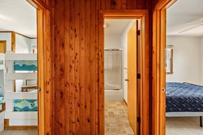 Ein Holzinterieur mit einer sichtbaren Tür zu einem Badezimmer, einem Bett mit gesteppter Bettdecke und einem weißen Regal mit aufgemaltem Dekor.