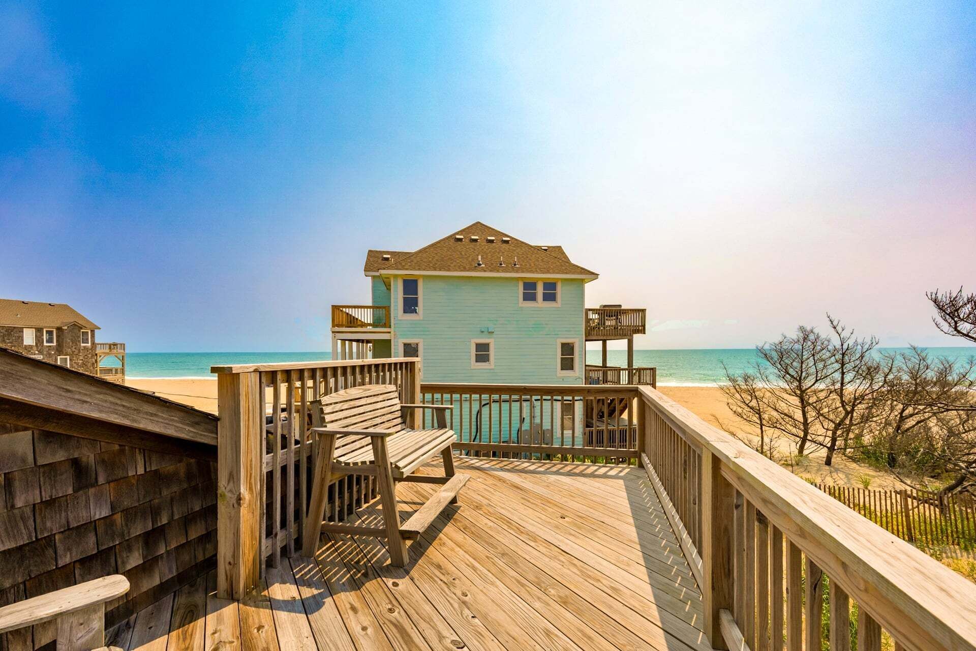 A wooden deck leading to a light blue house with a balcony, overlooking a beach.