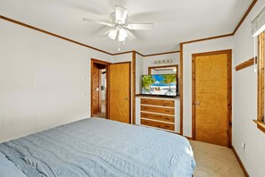 Een slaapkamer met een bed, een plafondventilator, een televisie op een houten kast en een deur die naar een andere kamer leidt.