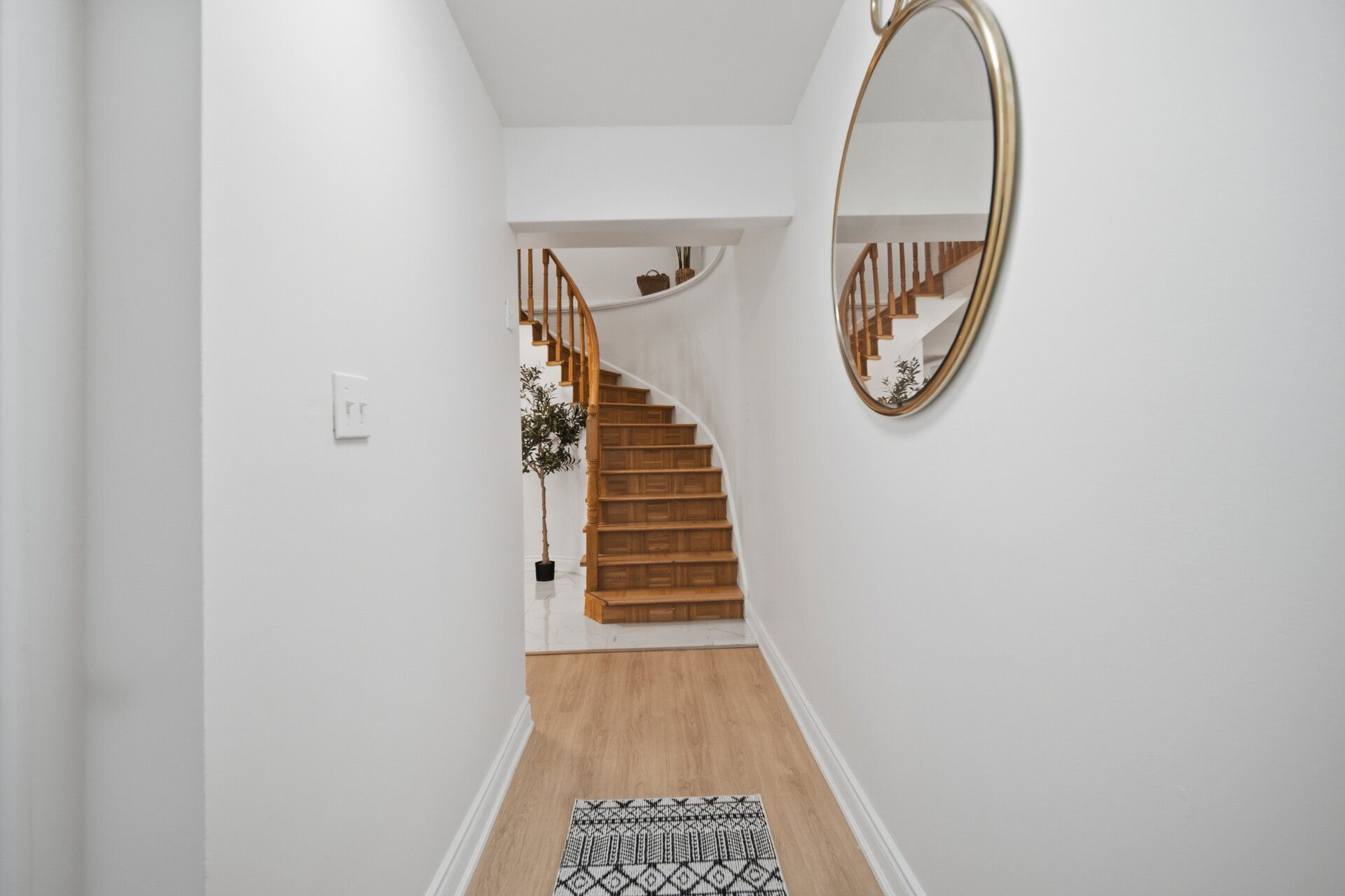 A hallway with a wooden staircase, a white wall, a mirror, and a patterned rug.