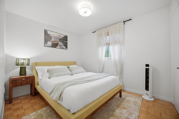 A bedroom with a bed, a bedside table with a lamp, a radiator, and a window with curtains.