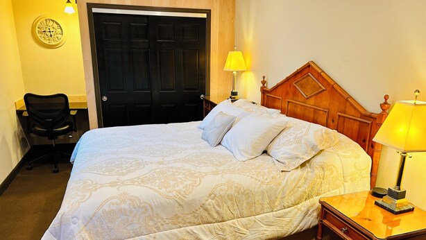 A hotel room with a large bed, a wooden headboard, a desk with a chair, and a door. - High end 4-bedroom countryside home in Shawano, WI. 2,800 sqft and spacious! (Shawano)