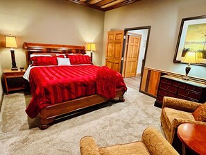 Une chambre à coucher avec un grand lit, une tête de lit en bois, deux tables de chevet avec des lampes, une commode et une porte donnant sur une autre pièce.