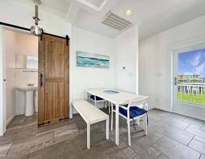 A room with a white table, bench, and chairs, a wooden door, a painting, and a view through a glass door. - Beach FRONT | 180 degree VIEWS | REVIEWS!!! (Crystal Beach)