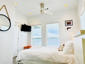 A bedroom with a bed, a ceiling fan, a TV, and two windows with blinds. - Beachfront Home w/ 180 Degree Views | Private (Crystal Beach)