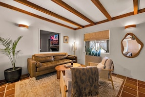 A living room with a leather sofa, a coffee table, a patterned rug, a potted plant, and a television displaying a desert landscape. - Boulder Oasis - 3 Bed 2 Bath Home in Yucca Valley (Yucca Valley)