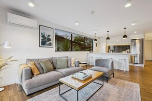 A modern living room with a grey sofa, a coffee table, and a kitchen area with a white countertop and stainless steel appliances. - Queenstown Quarters - Residence I (Queenstown)