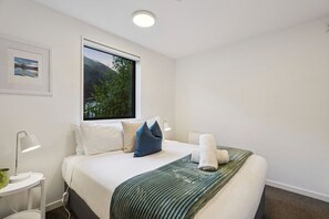 A hotel room with a bed, a window with a view of greenery, a bedside table with a lamp, and a framed picture on the wall. - Queenstown Quarters - Residence I (Queenstown)