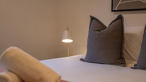 Una cama bien hecha con almohadas, una lámpara de noche y una toalla enrollada sobre una mesita de madera.