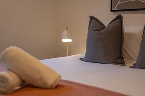Ein ordentlich bezogenes Bett mit Kissen, einer Nachttischlampe und einem aufgerollten Handtuch auf einem Holznachttisch.
