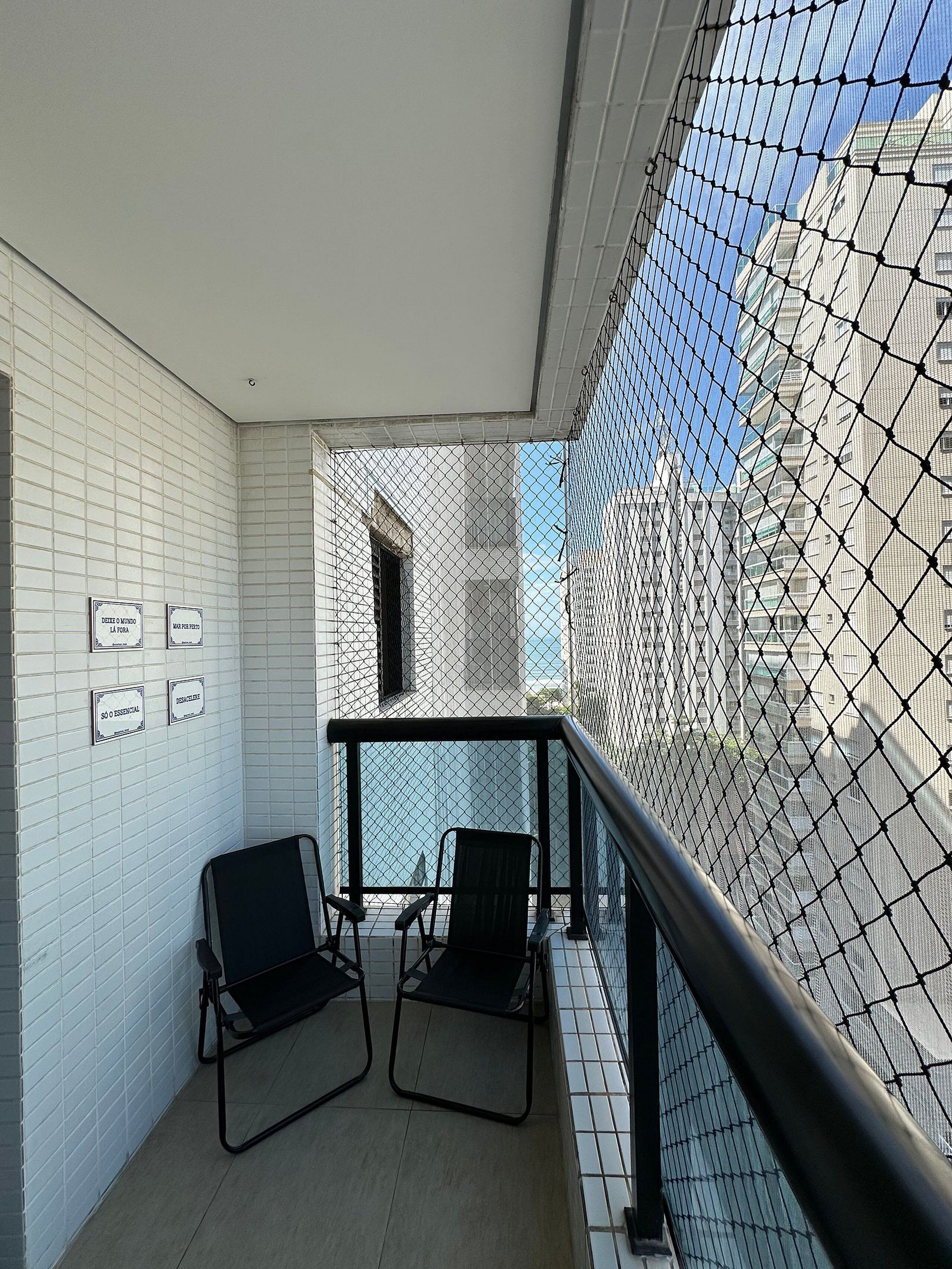 A balcony with two chairs, white tiled walls, and a view of buildings through a mesh screen.