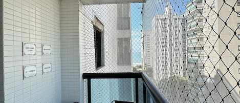 Un balcon avec deux chaises, des murs recouverts de carreaux blancs et une vue sur les immeubles à travers un moustiquaire.